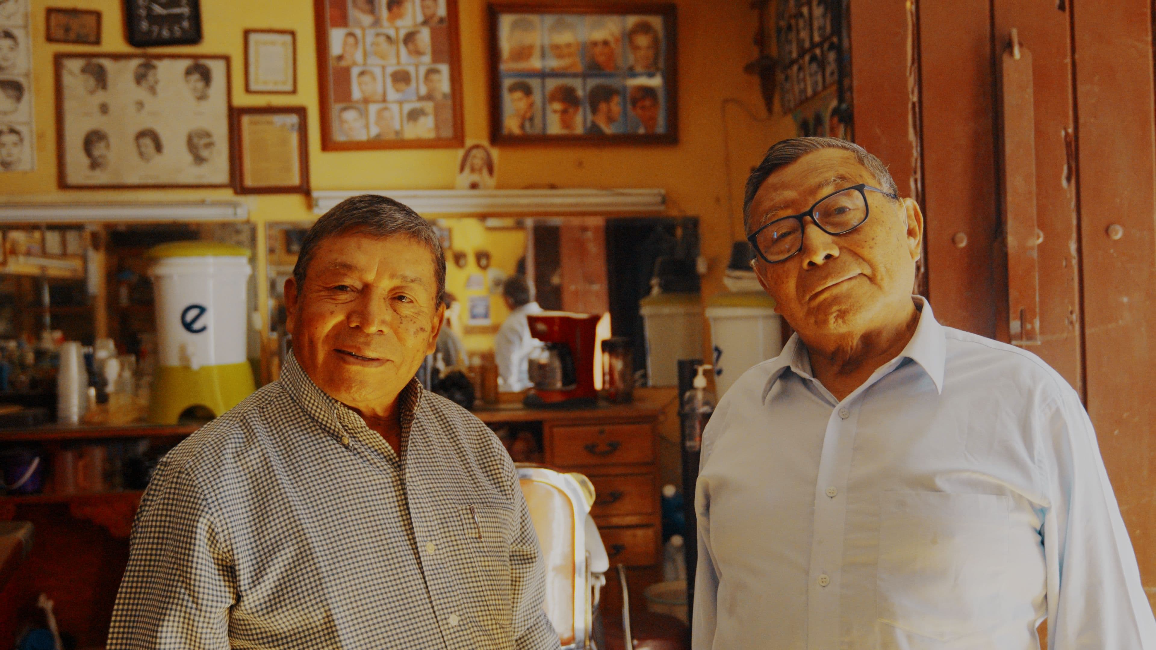 Two elderly barbers standing inside a traditional barbershop in Antigua Guatemala, surrounded by mirrors and hairstyle photos.